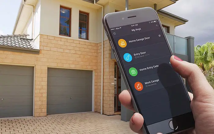 Mr. Goldie Garage Door - Hand holding a smartphone showing a smart home app with four door controls in front of a brick house with two garage doors stained orange.