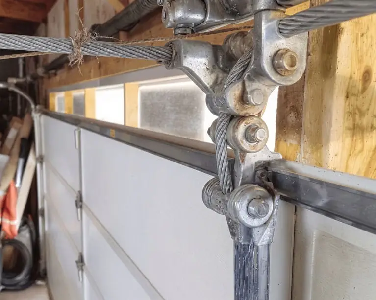 Mr. Goldie Garage Door - Mr. Goldie Garage Door - Close-up of a garage door torsion spring and steel cables attached to a metal bracket and track hardware in a garage.